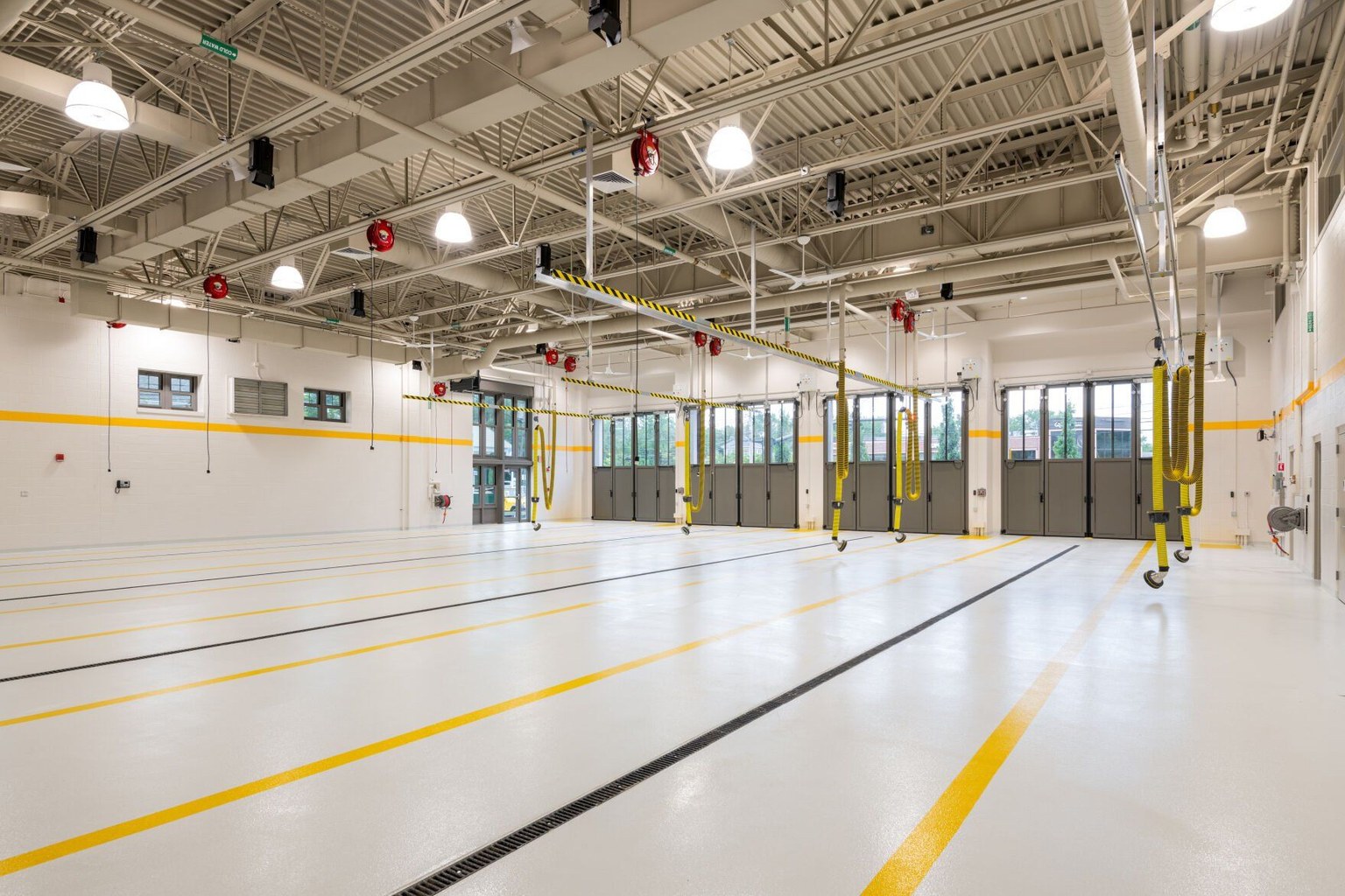 Empty fire station apparatus bays with a light finished floor, yellow lane lines, and suspended exhaust hoses.