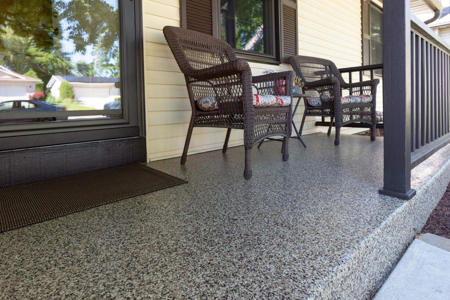 Covered front porch with a speckled coating, wicker chairs, yellow siding, and dark railing.